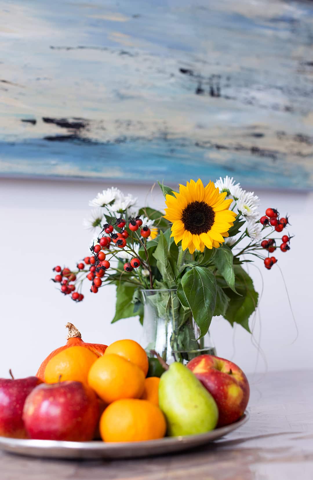 Blumenstrauß mit Sonnenblume und Obstteller auf Tisch vor ruhigem, blauem Gemälde im Hintergrund