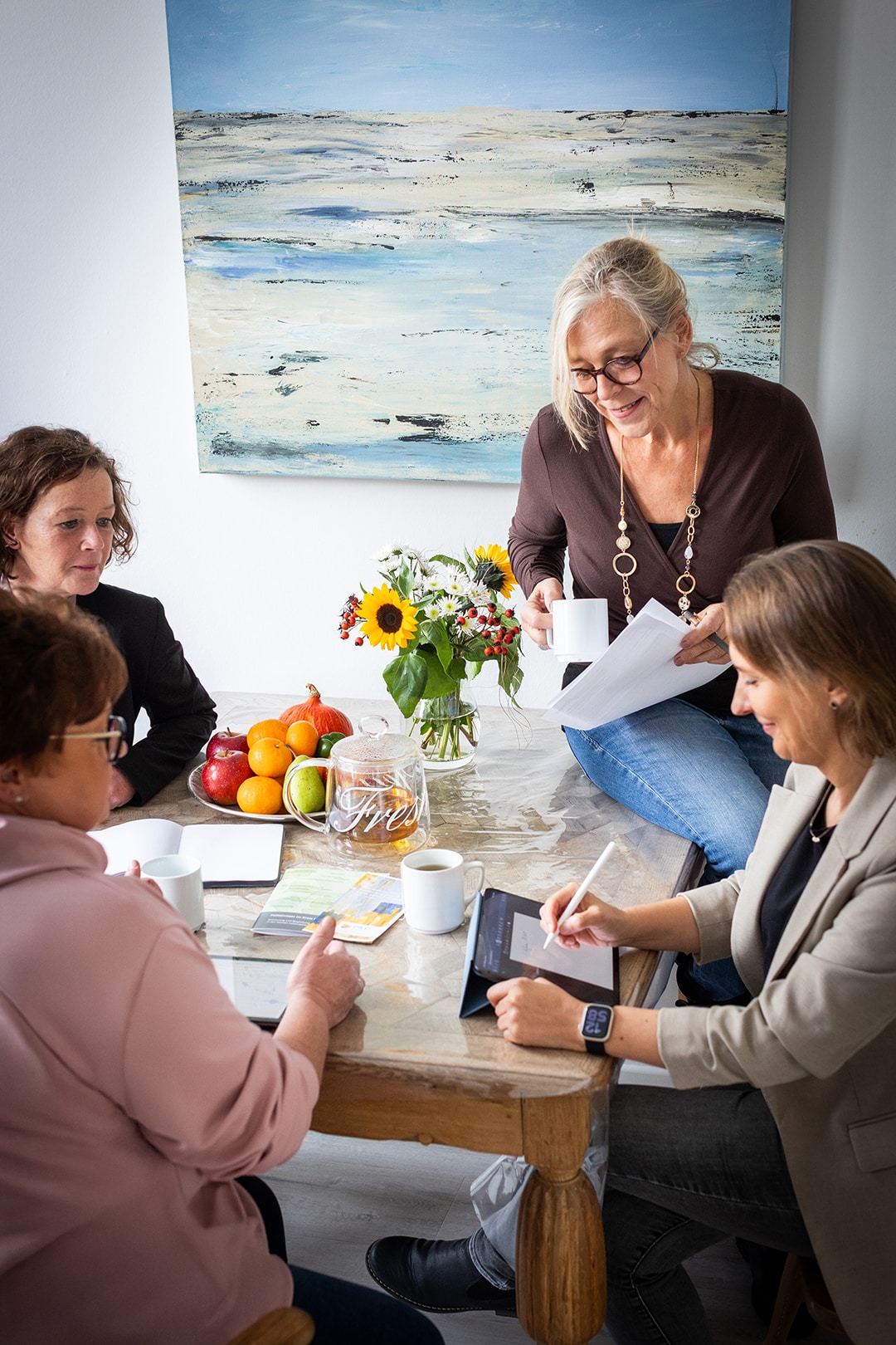 Pflegedienst-Team beim Gespräch um einen Tisch, umgeben von Blumen und Obstschale in häuslicher Atmosphäre
