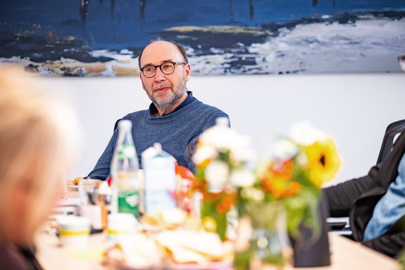 Einfaches Gespräch am Tisch in ruhiger häuslicher Umgebung, im Fokus eine pflegende Fachkraft mit Blumen im Vordergrund